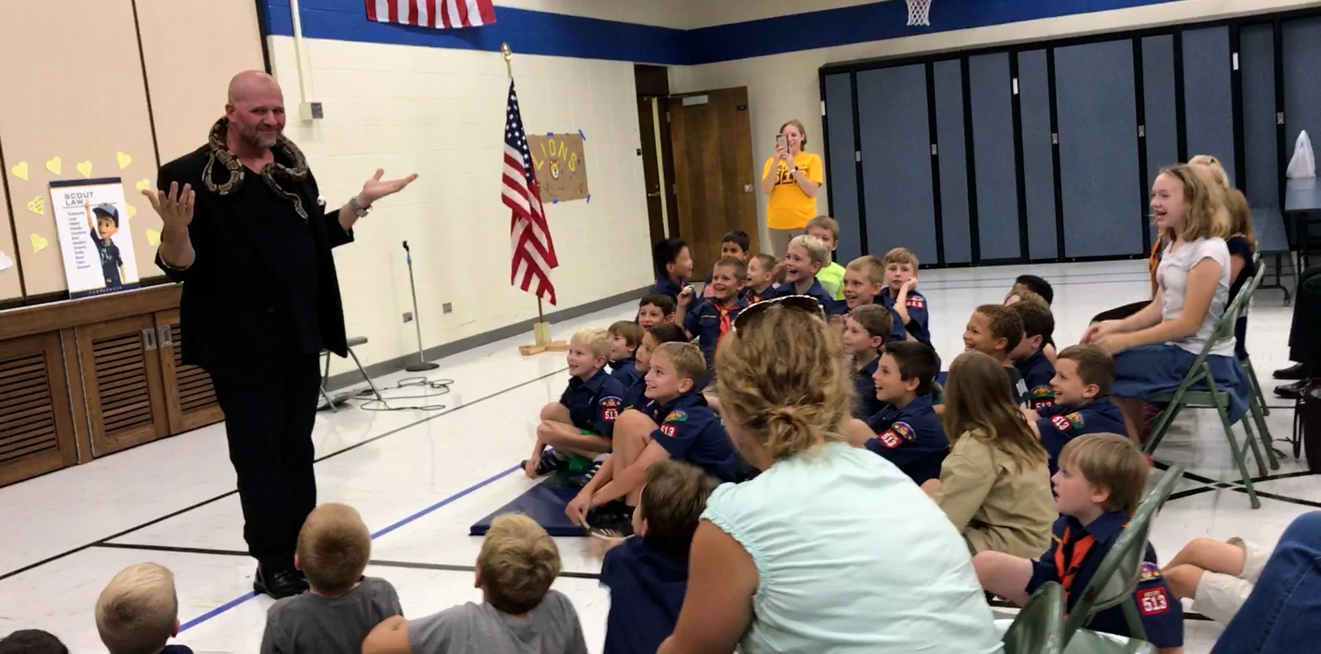 Mike DiDomenico with python around his neck performing for Pack 513 Scouts with American flag and Scout Law poster visible
